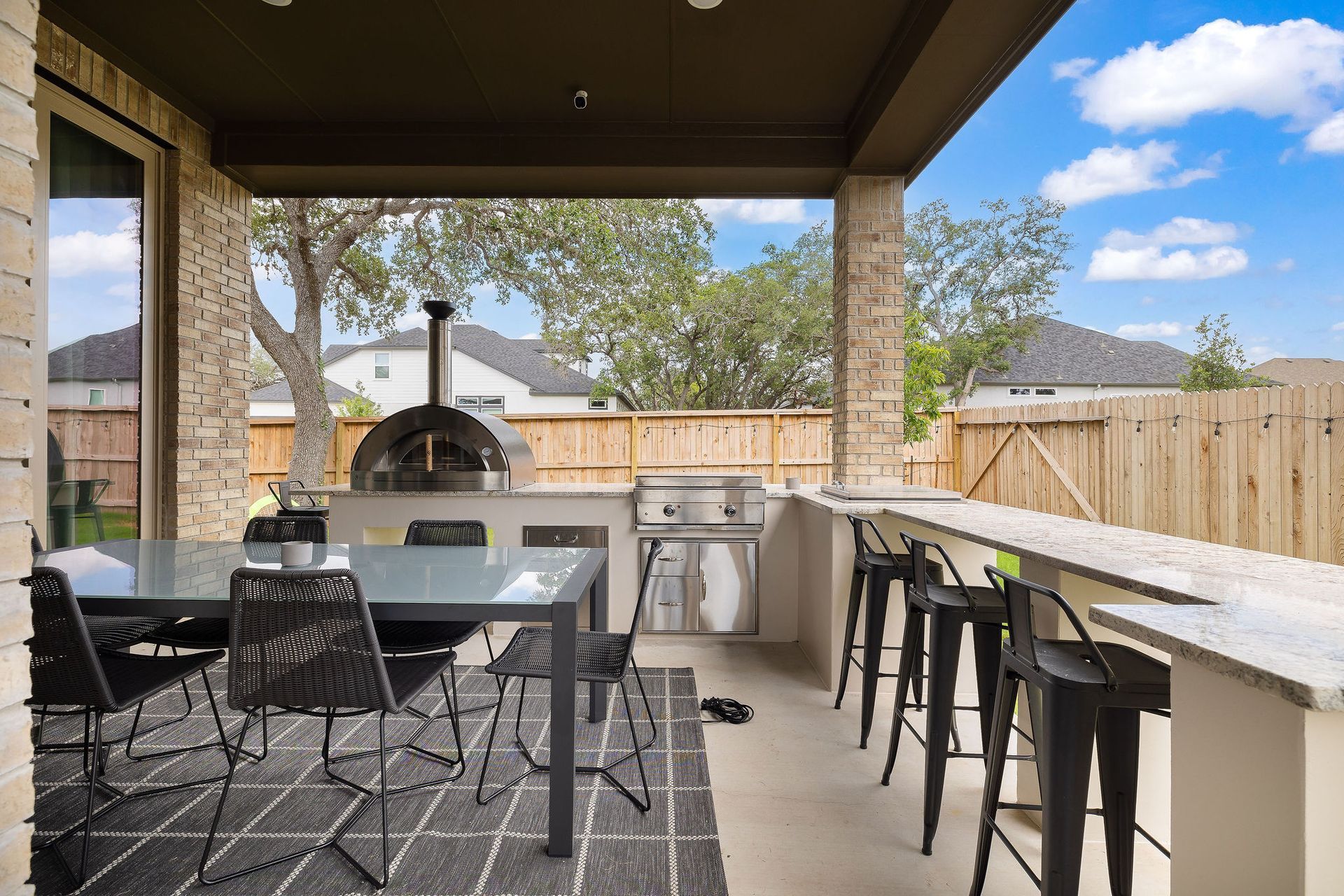Covered patio with dining table, bar seating, and grill overlooking a fenced backyard under a blue sky