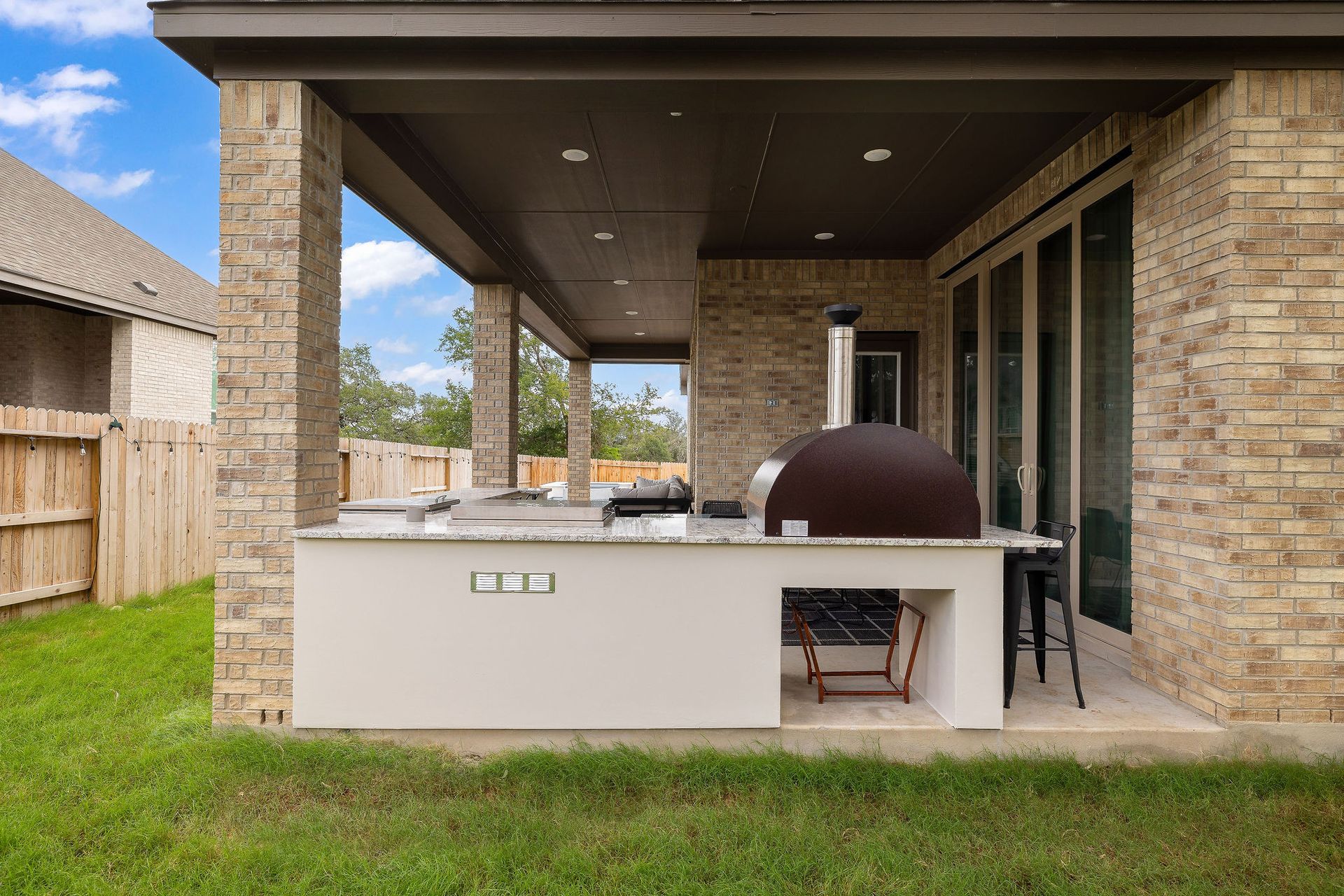 Covered patio with outdoor kitchen island, built-in grill, and brick house exterior beside a grassy yard
