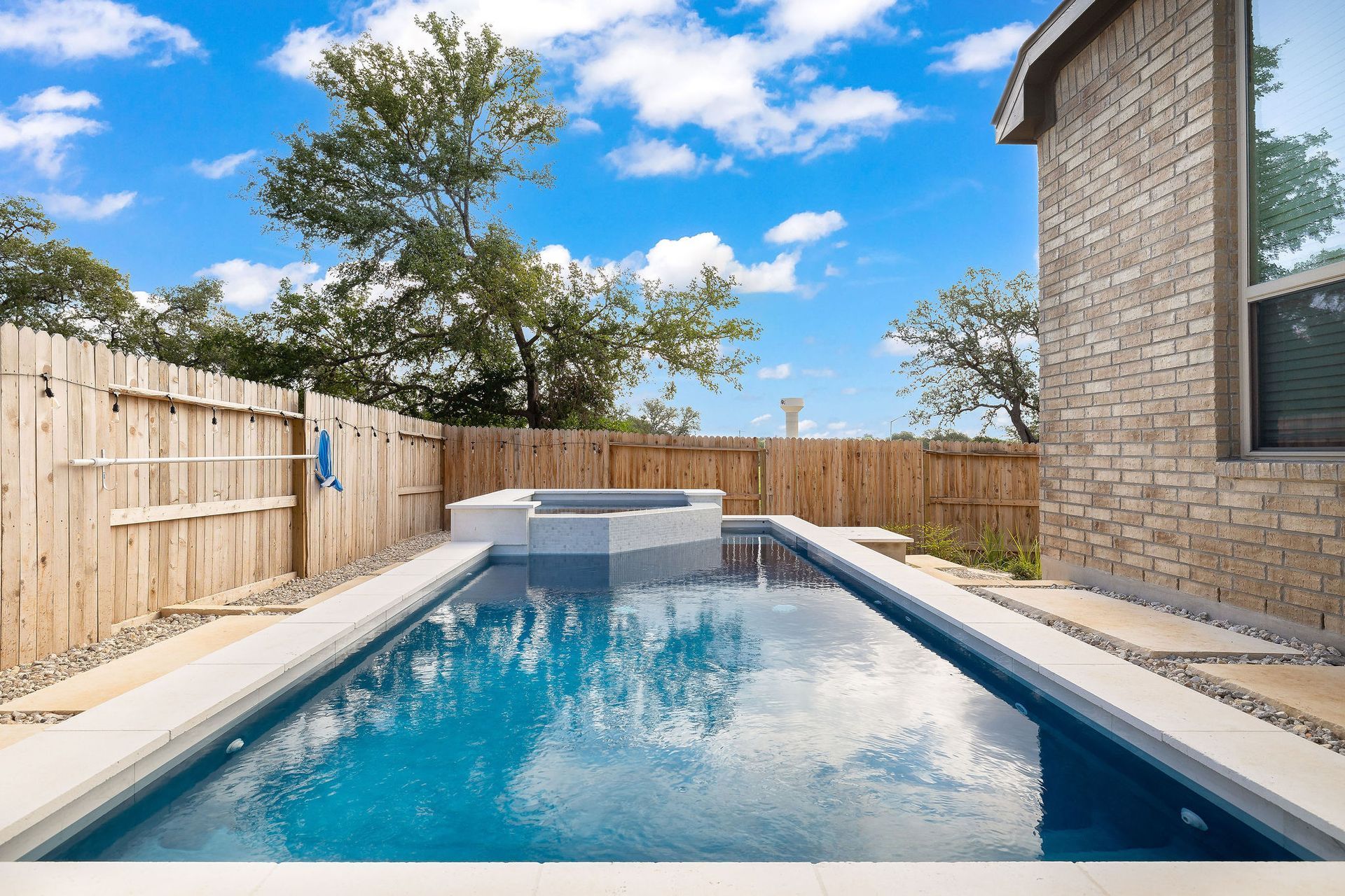 Rectangular backyard swimming pool beside a brick house and wooden fence under a blue sky with clouds