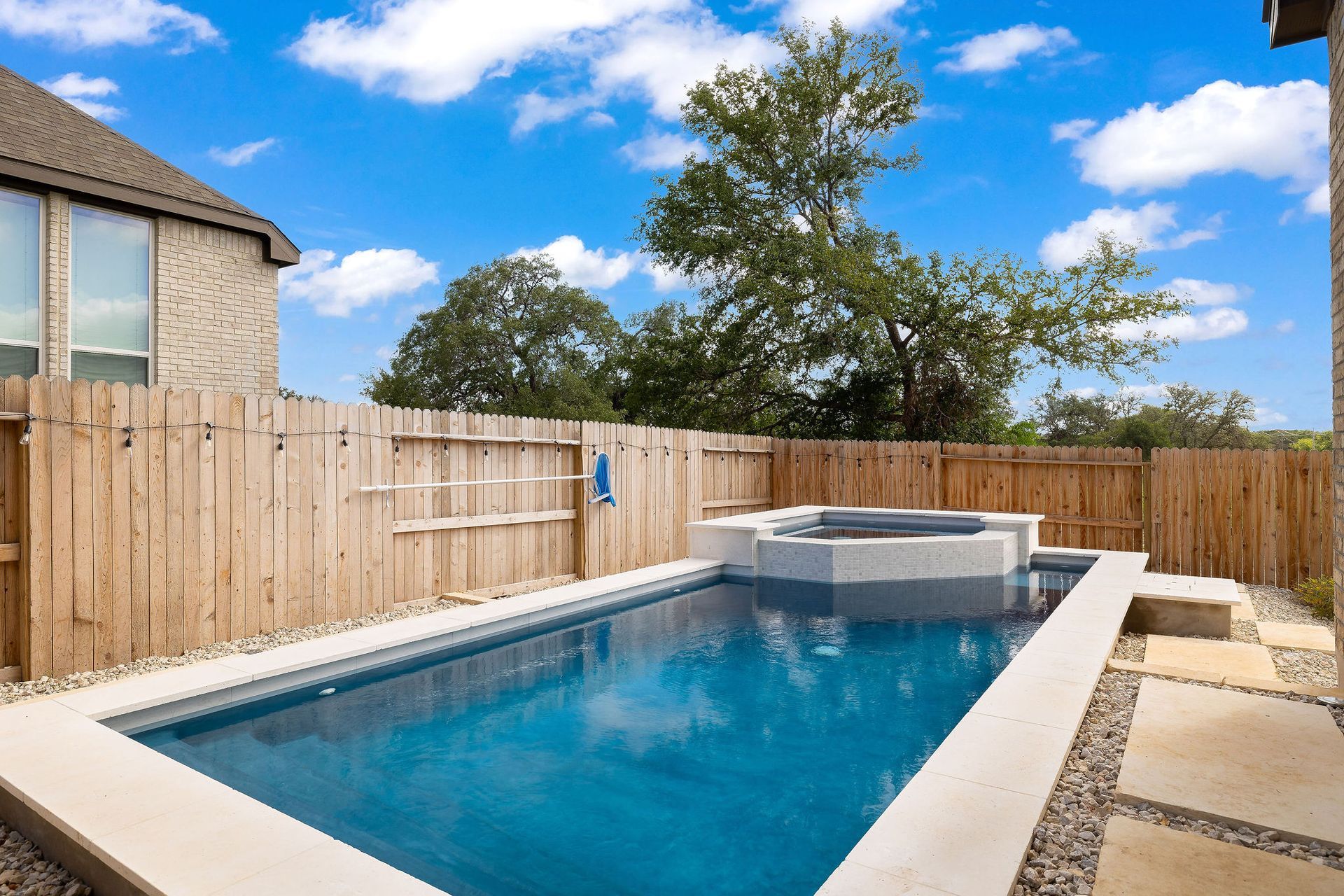 Backyard swimming pool with a raised spa, wooden fence, and blue sky.
