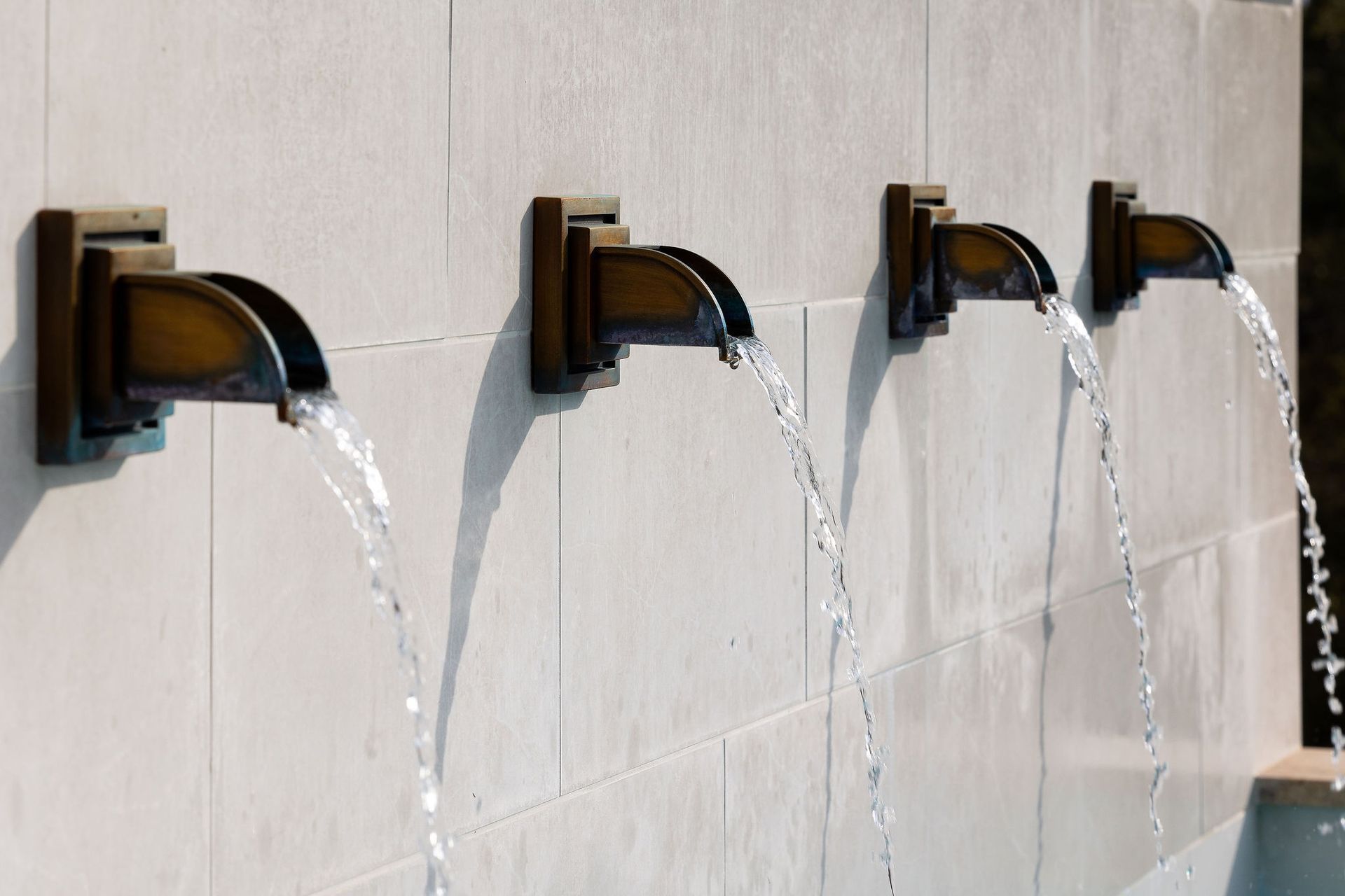 Four wall-mounted faucets pour water into sinks against a white tiled wall.