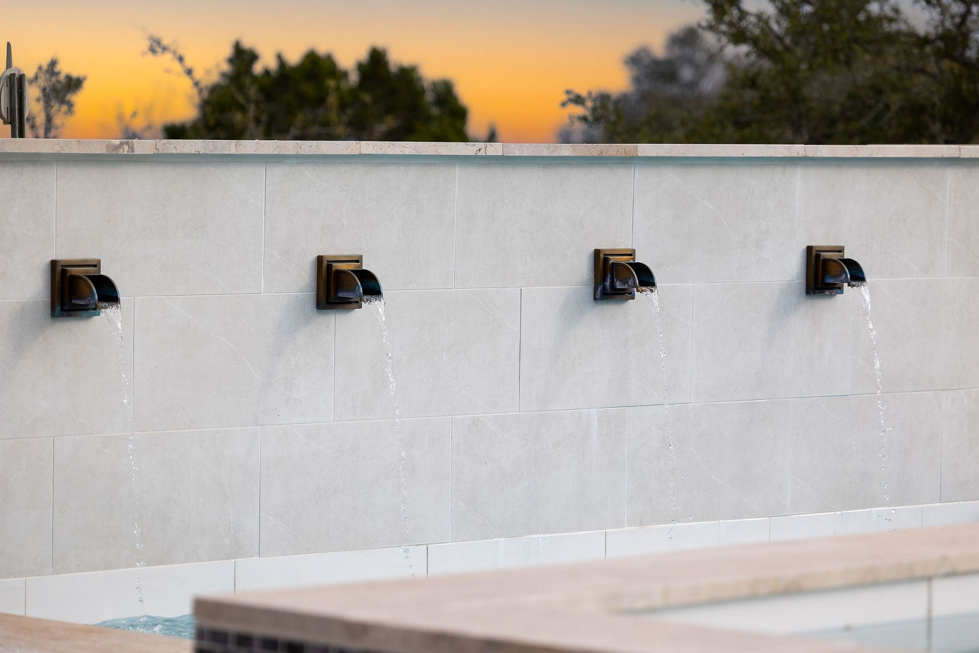 White tiled wall with four black faucets at sunset, with trees in the background.