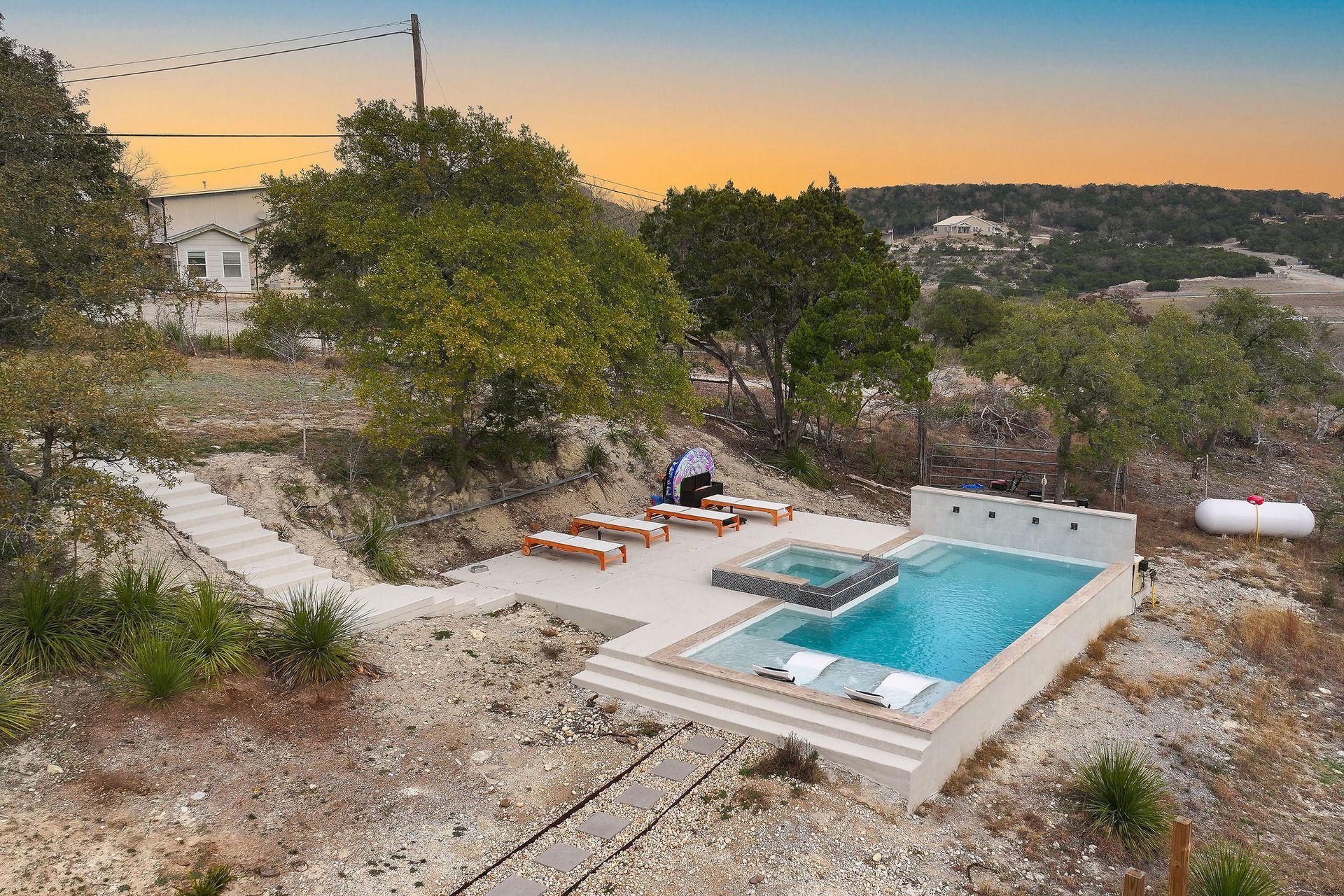 Modern backyard with a turquoise pool, lounge chairs, trees, and a sunset sky over hills.