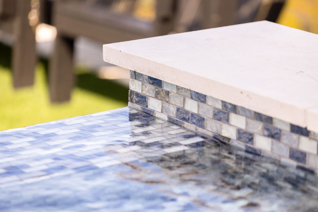 Blue mosaic-tiled pool edge with a white coping and blurred lawn in the background
