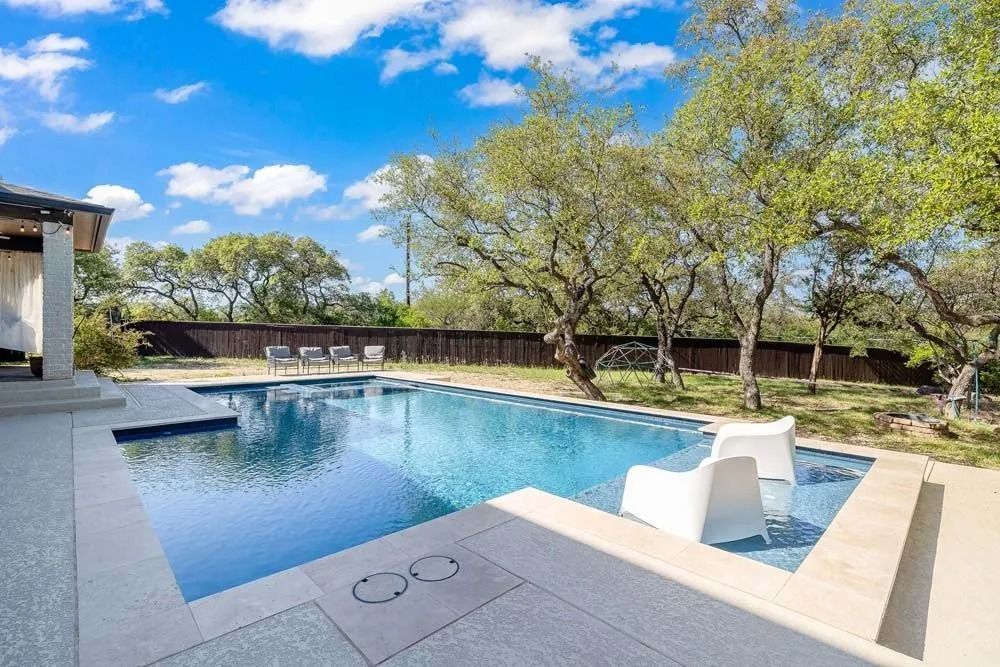 A backyard swimming pool with a sun shelf and two white chairs, set against a backdrop of trees and a bright blue sky.