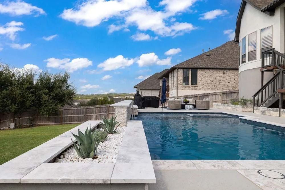 A backyard swimming pool with light-colored stone coping, a planter box with agave plants, and a house in the background.