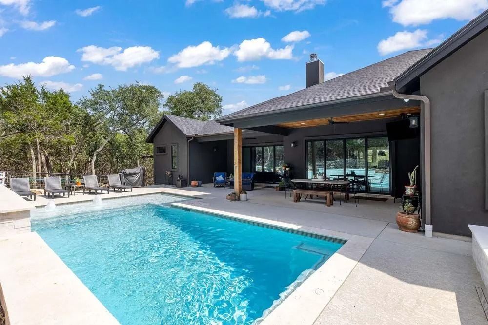 A backyard swimming pool with a modern dark grey house, covered patio, and lounge chairs on a sunny day.