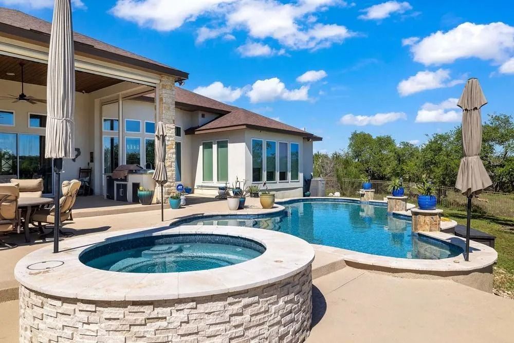 A patio with a circular hot tub and a swimming pool overlooking a backyard with trees under a bright blue sky.