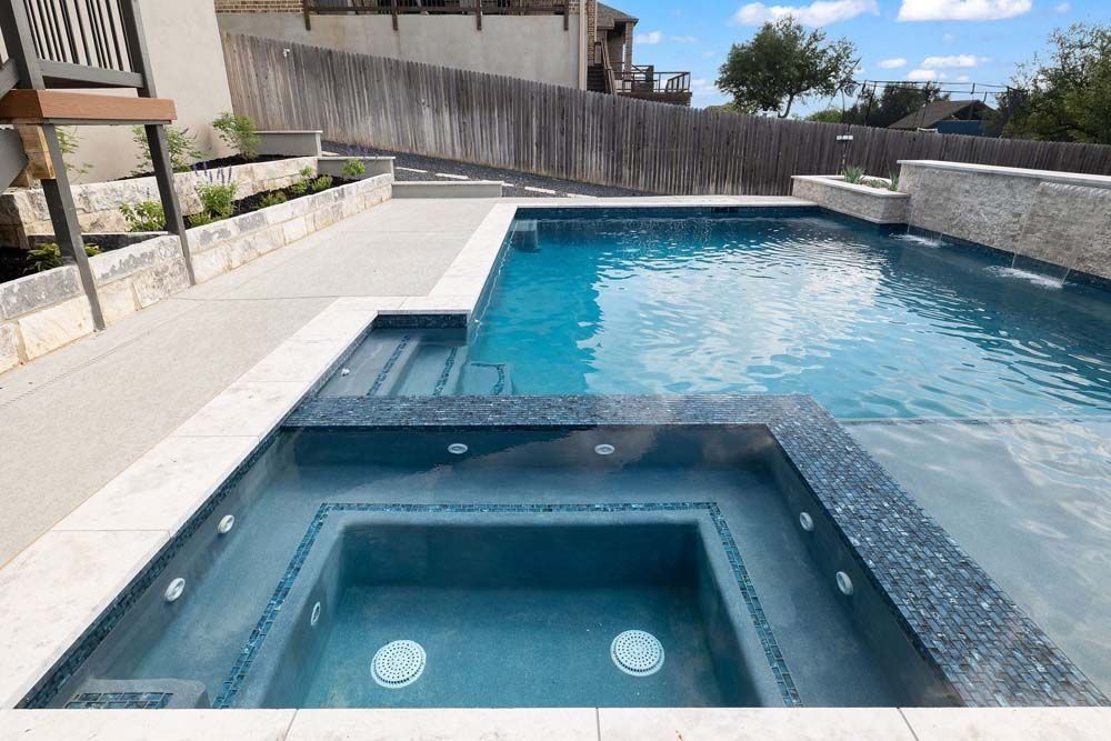 A bright blue swimming pool with an attached square spa, featuring light stone trim and a concrete pool deck.