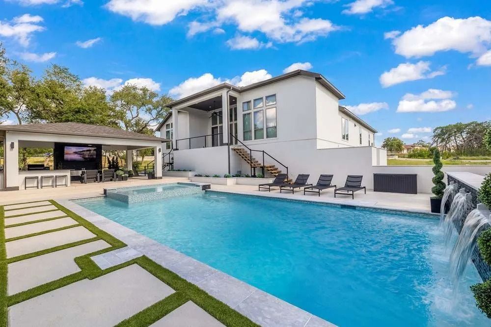 A bright, modern house with a pool, spa, waterfall feature, outdoor covered seating area, and grass-paved deck.