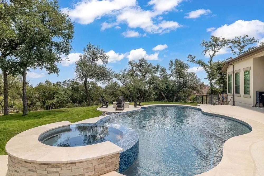 A free-form swimming pool with a circular spa and stone accents in a backyard setting with trees and a blue sky.