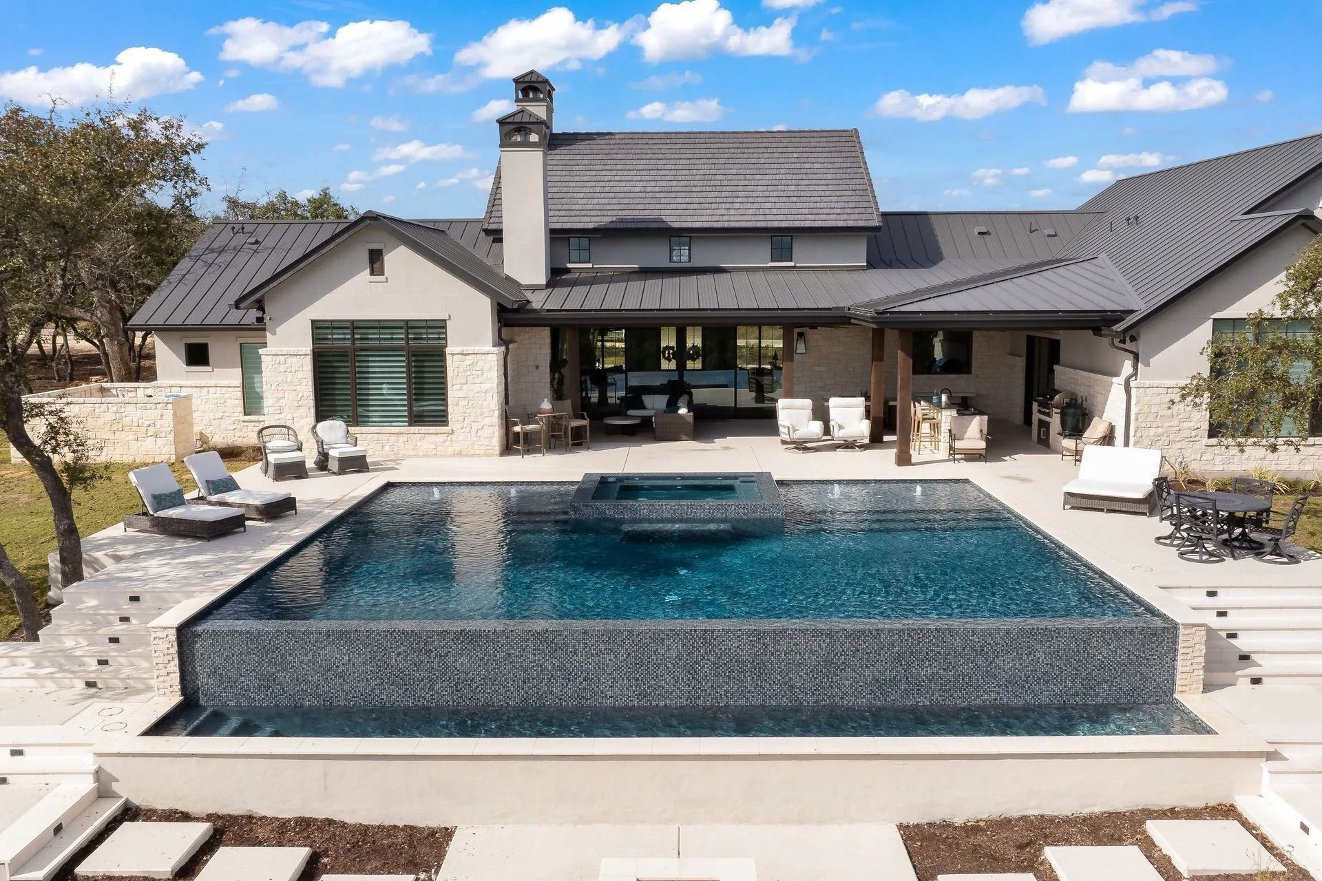 A modern house with a dark metal roof, stone exterior, and a large infinity pool with a built-in spa in the backyard.