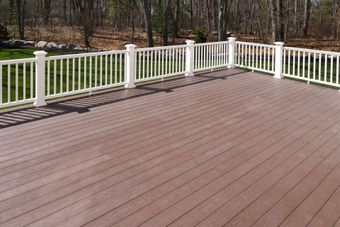 A wooden deck with a white railing overlooks a green backyard and trees on a sunny day.