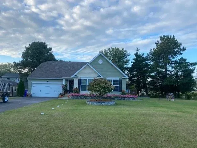 Suburban house with a gray roof, white garage, flower beds, and a large front lawn under a cloudy sky