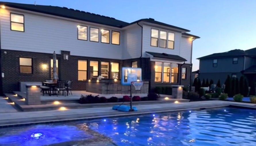 Backyard pool and patio at twilight, featuring modern two-story home, lit landscape, and basketball hoop.