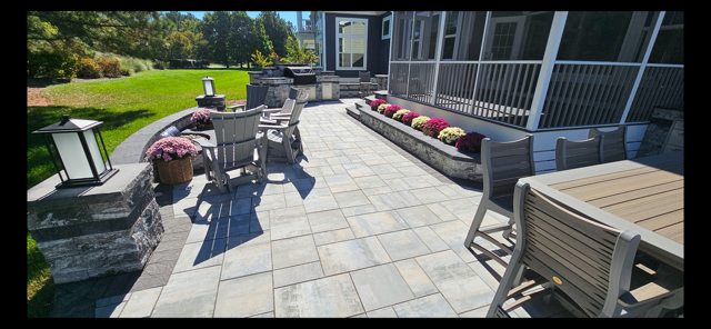 Sunny patio with outdoor dining tables, lounge chairs, potted flowers, and a grassy yard beyond.