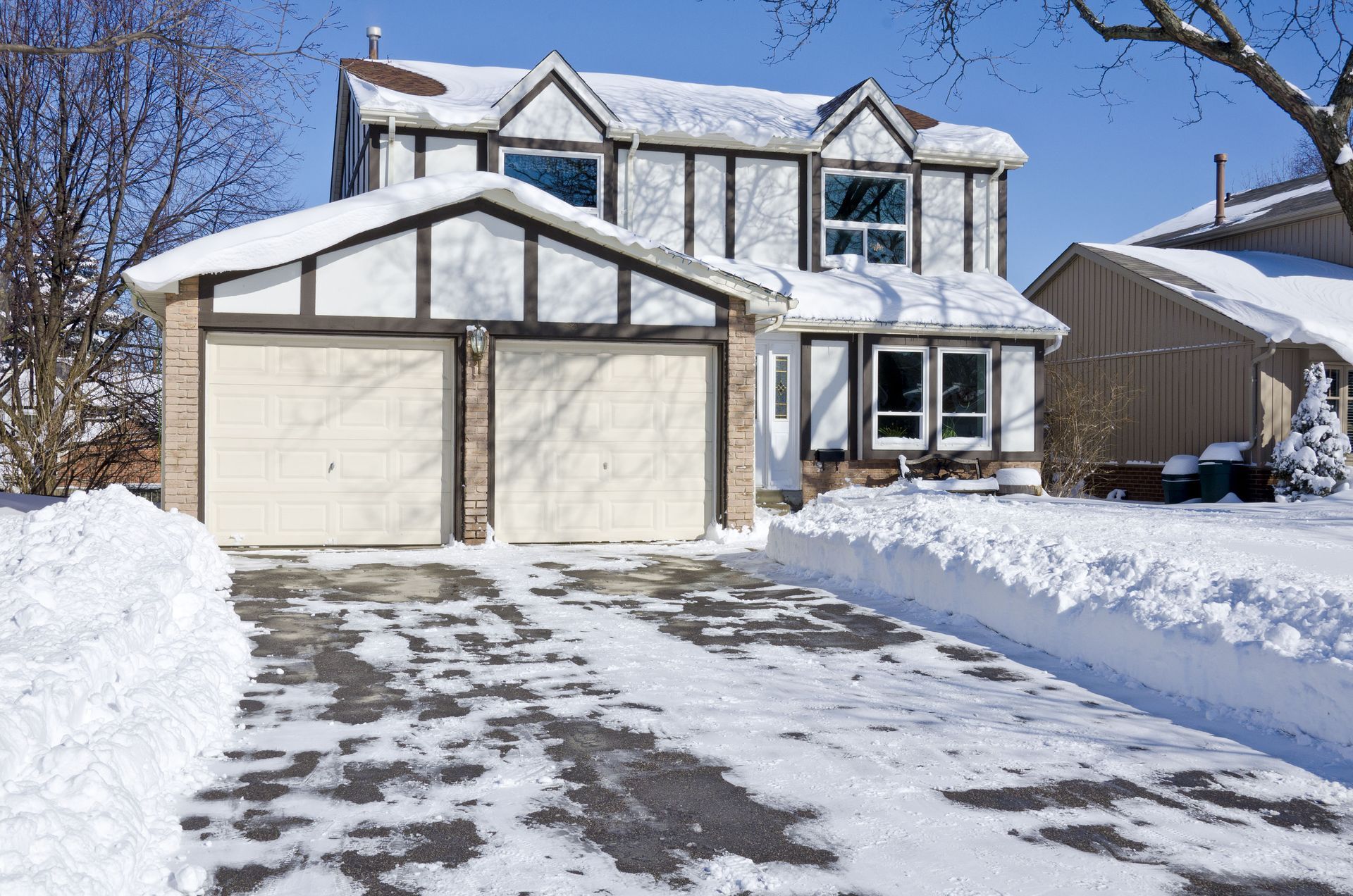 Two-story house with attached garage covered in snow on a sunny winter day. Driveway has melted patches.