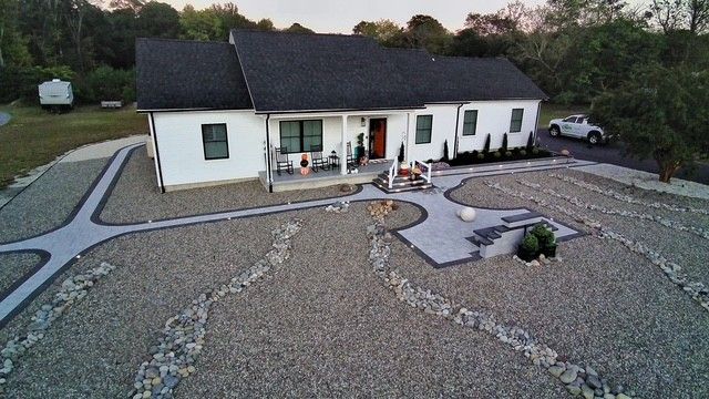 White house with dark roof, front porch, and stone landscaping. Gravel yard with walkway and car.