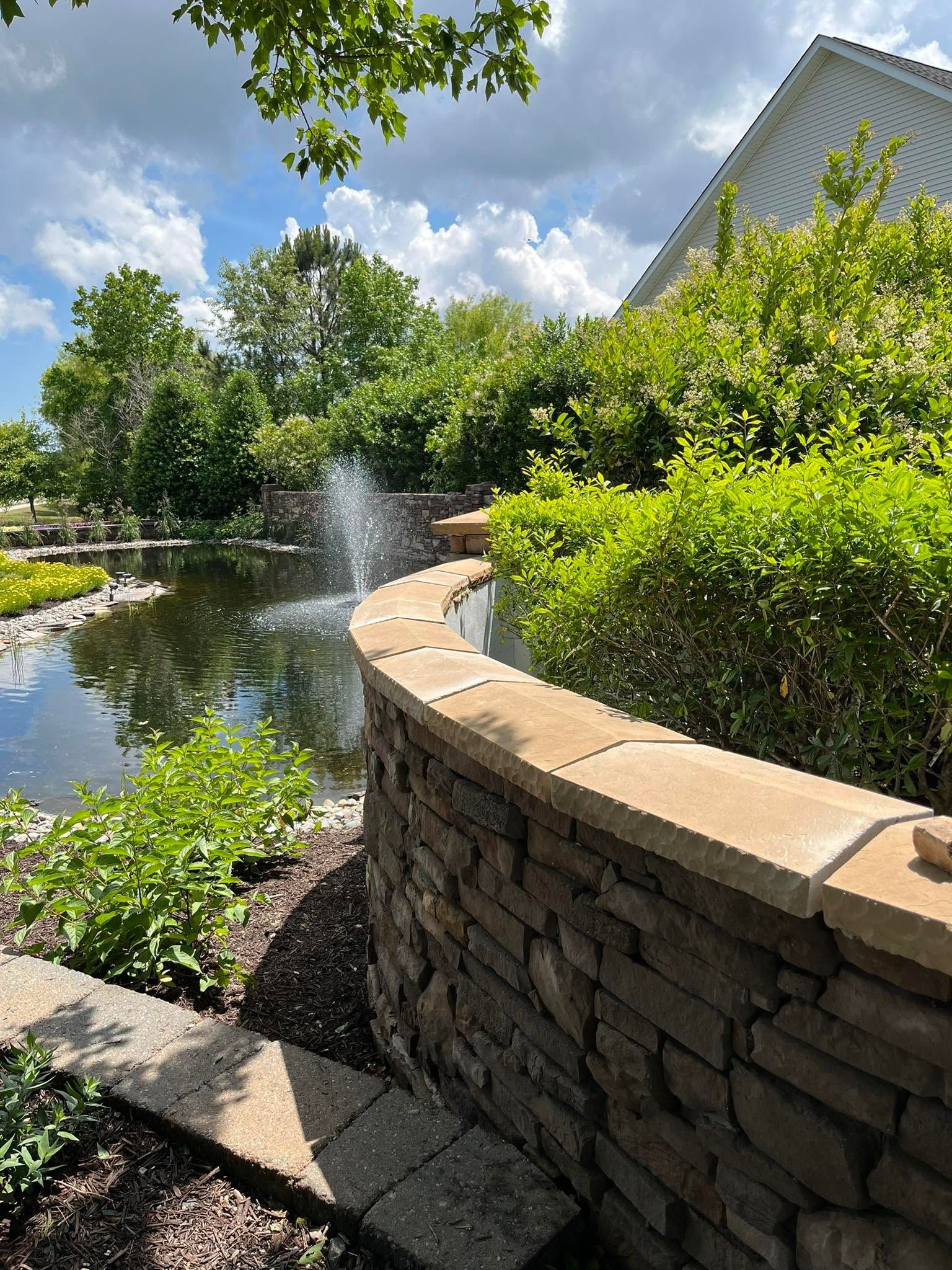 A stone wall surrounds a pond with a fountain in the background.