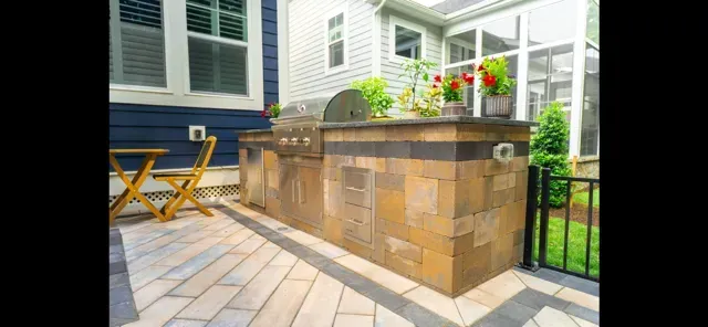 Patio with tan stone bar, black railing, potted flowers, and wooden chairs beside a house