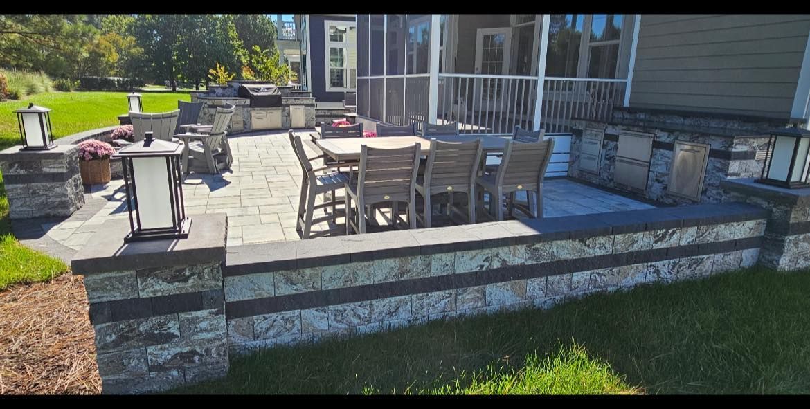Patio with outdoor dining table, chairs, and grill beside a house, bordered by a stone retaining wall.