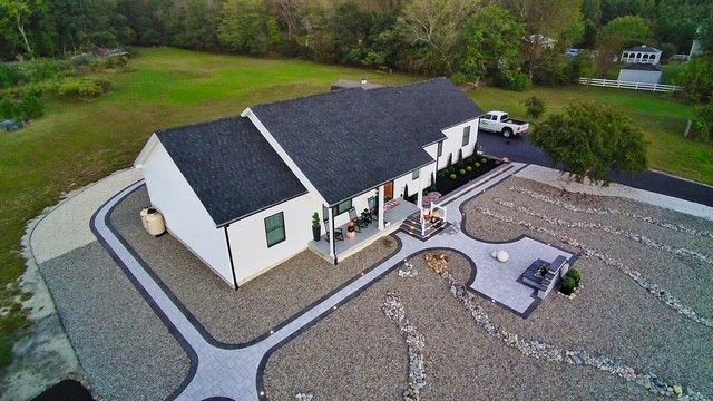 Aerial view of a white house with a dark roof, gravel yard, and patio seating surrounded by greenery.