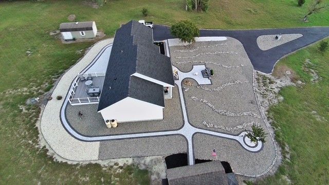 Aerial view of a house with a dark roof, gravel yard, curved paths, and a small driveway.