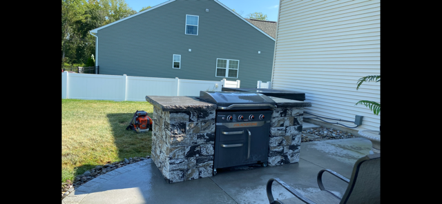 Outdoor stone grill on a patio beside a house and fenced yard