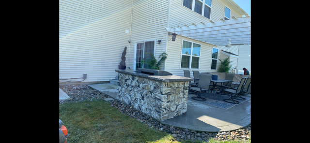Stone patio with a built-in grill and pergola beside a beige house, with outdoor seating nearby.
