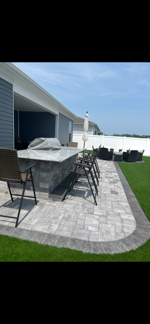 Patio dining area with gray table and chairs beside a house, overlooking a green lawn and white fence.