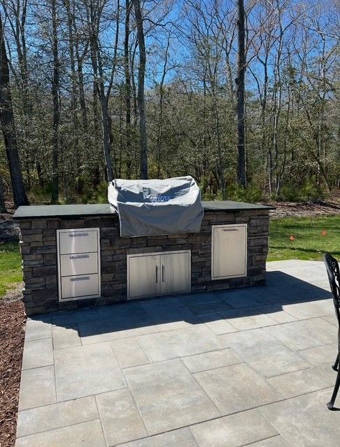 Backyard outdoor kitchen with stone cabinets and a covered grill on a patio, trees in the background