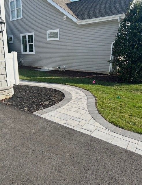 Curved stone walkway beside a gray house, bordered by grass and a dark mulch bed.
