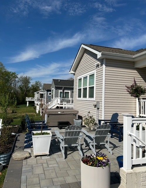 Patio with gray chairs, potted flowers, grill, and beige house under a blue sky