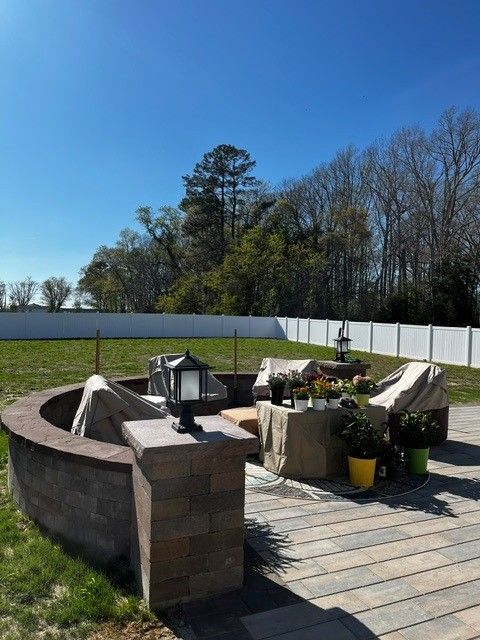 Backyard stone fire pit with lanterns and potted plants on a sunny lawn, white fence and trees behind