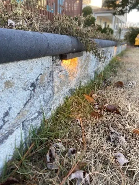 Stone retaining wall with black pipe and warm light, beside dry grass and fallen leaves.