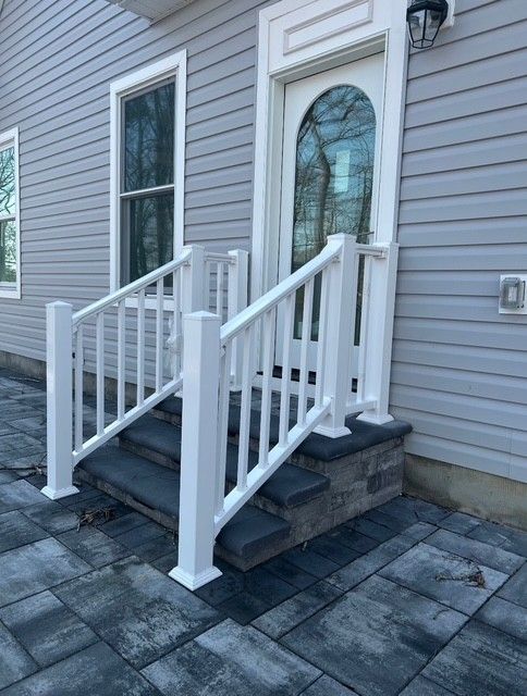 Front porch with white railings, dark steps, and stone pavers beside a gray house with a stained-glass door.