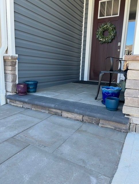 Front porch with gray siding, dark door with wreath, step, and several blue and purple flower pots.