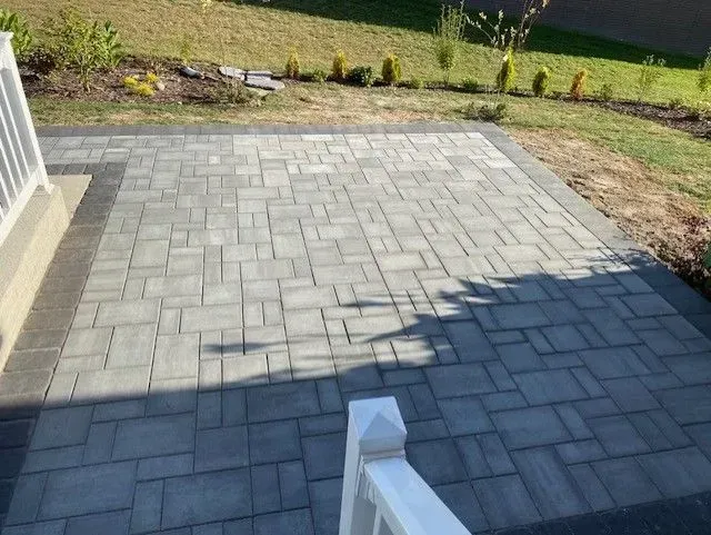 Paved stone patio with a small garden border and lawn in the background, partly shaded by the house.