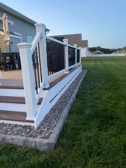 White deck railing beside a grassy yard at a house exterior, with stone edging and steps on the left