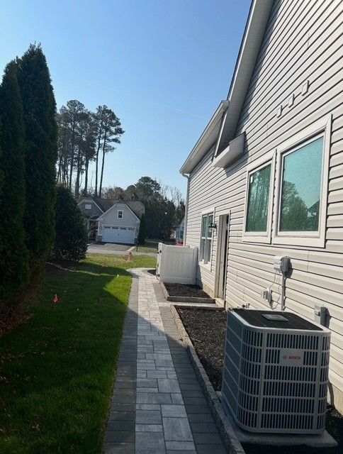 Side of a house with an HVAC unit, paved walkway, and grassy yard under a clear blue sky.
