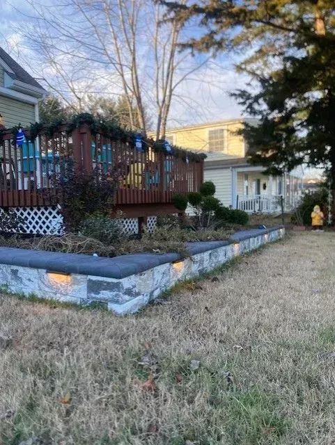 Backyard deck with holiday lights, stone retaining wall, and a white house beside a grassy yard