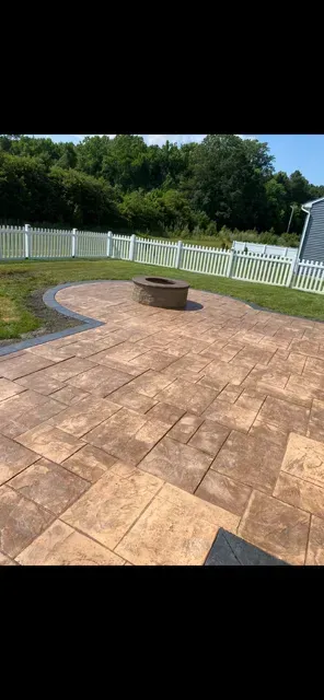 Stone patio with a round fire pit, surrounded by white fencing and green trees in bright sunlight