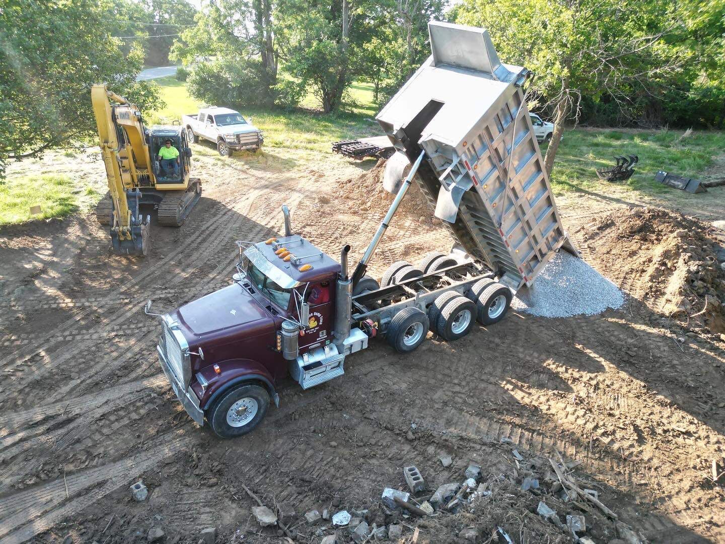Maroon dump truck unloading gravel at a construction site, with excavator and pickup nearby
