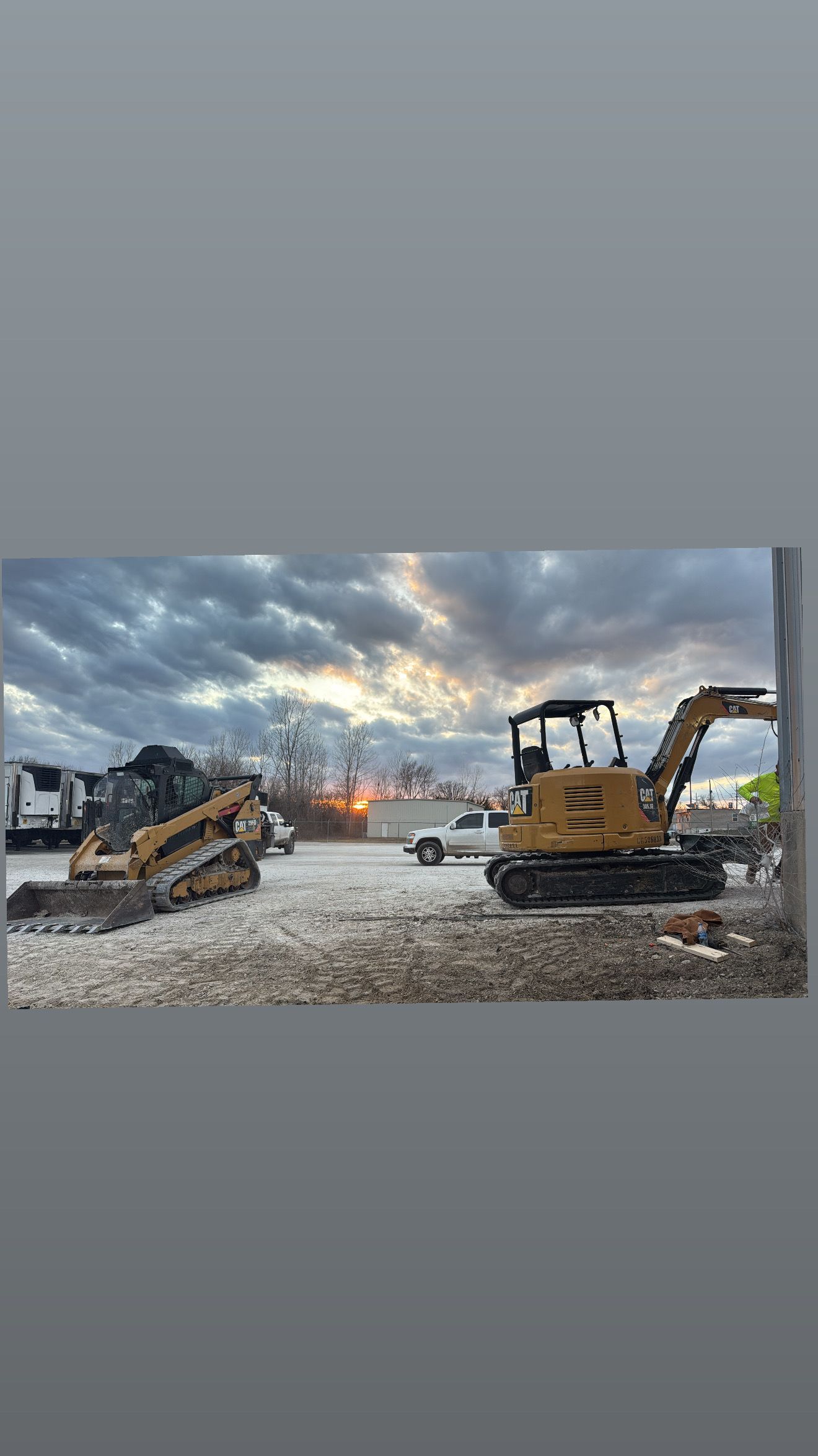 Two excavators on a gravel lot under a cloudy sunset sky
