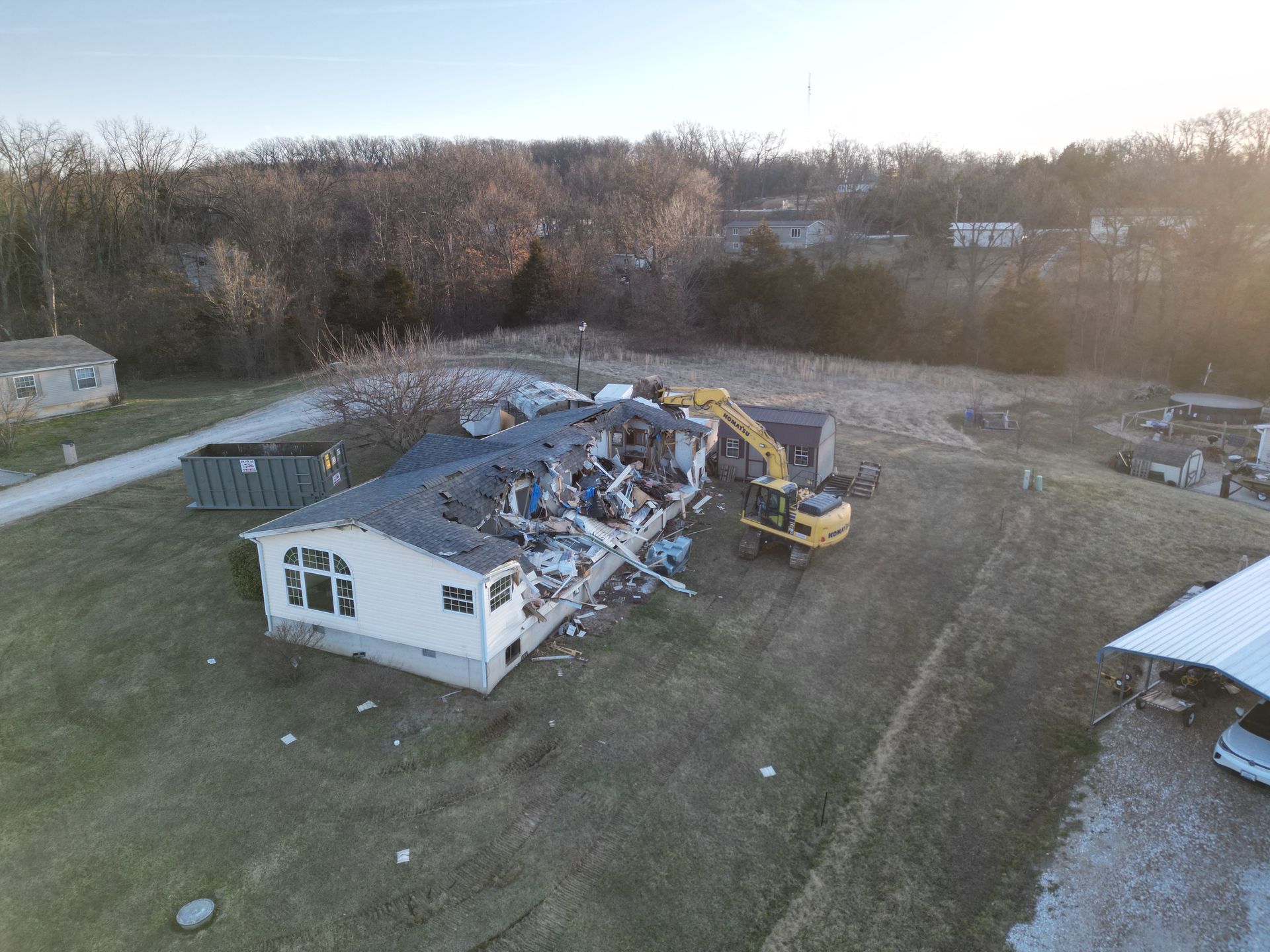 Yellow excavator demolishing a house in a rural field with debris scattered вокруг