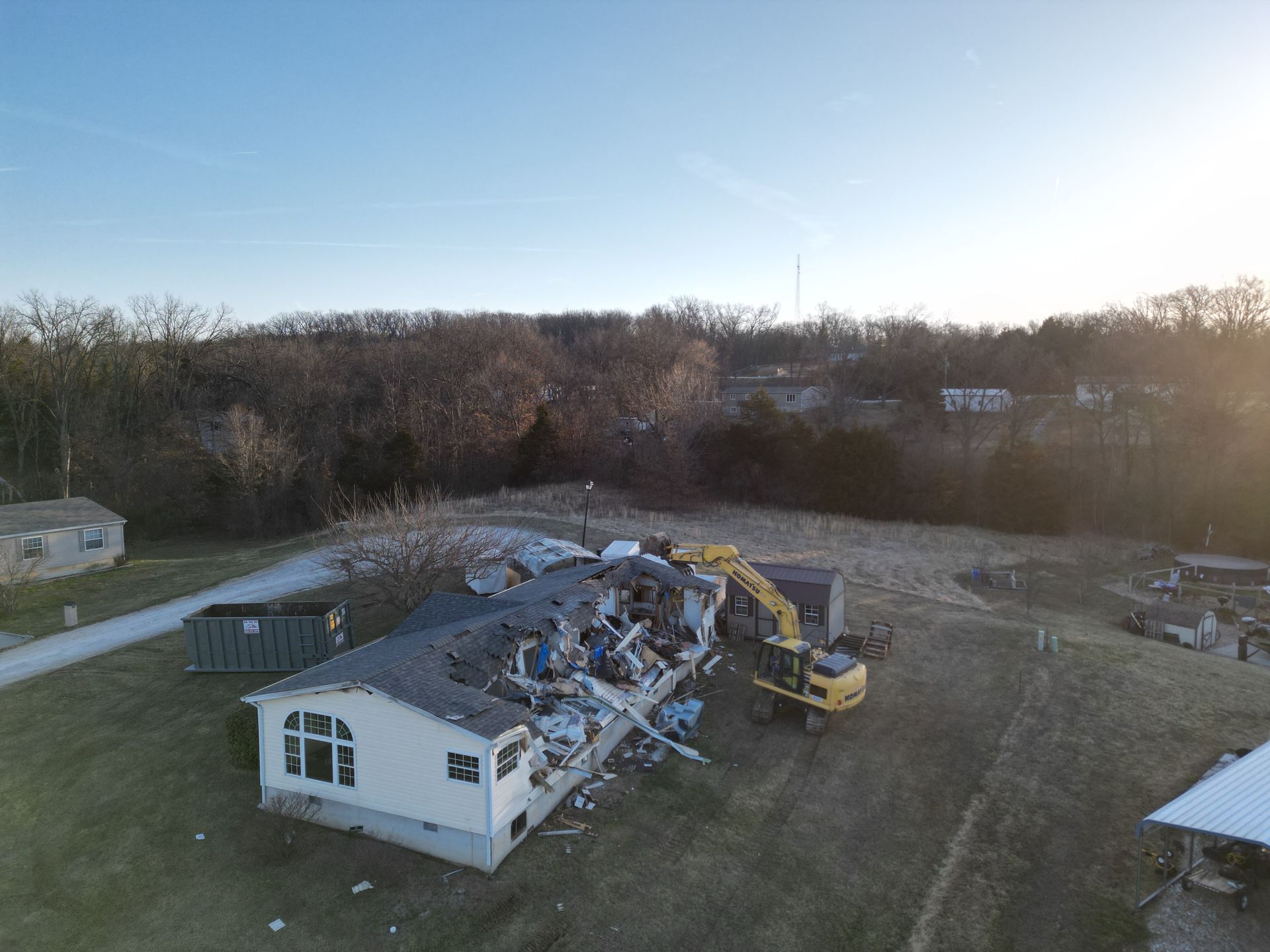 Damaged mobile home with collapsed roof, debris, and a yellow excavator in a wooded lot at sunset