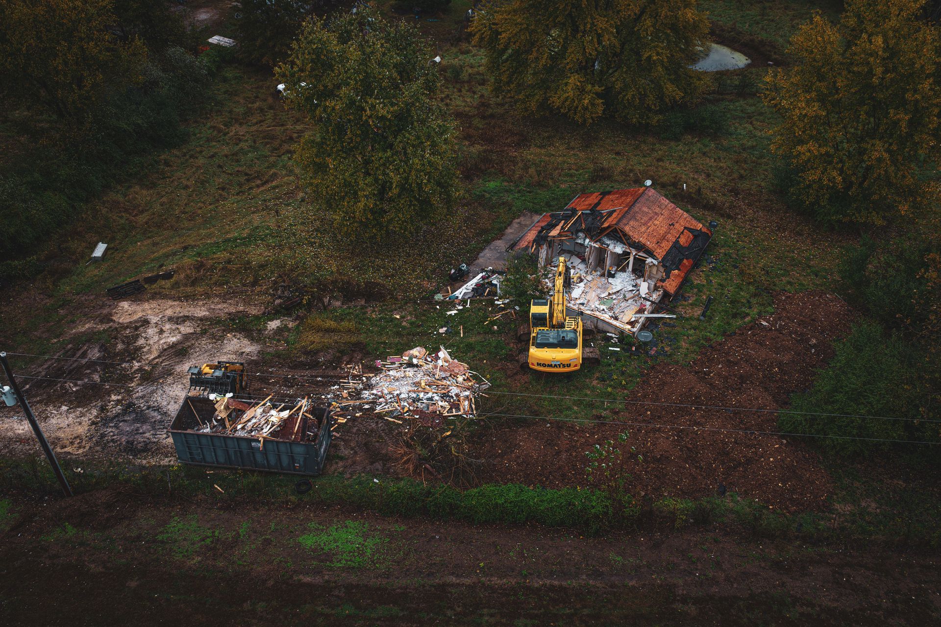 Aerial view of a yellow excavator beside a damaged small house and debris in a wooded area.