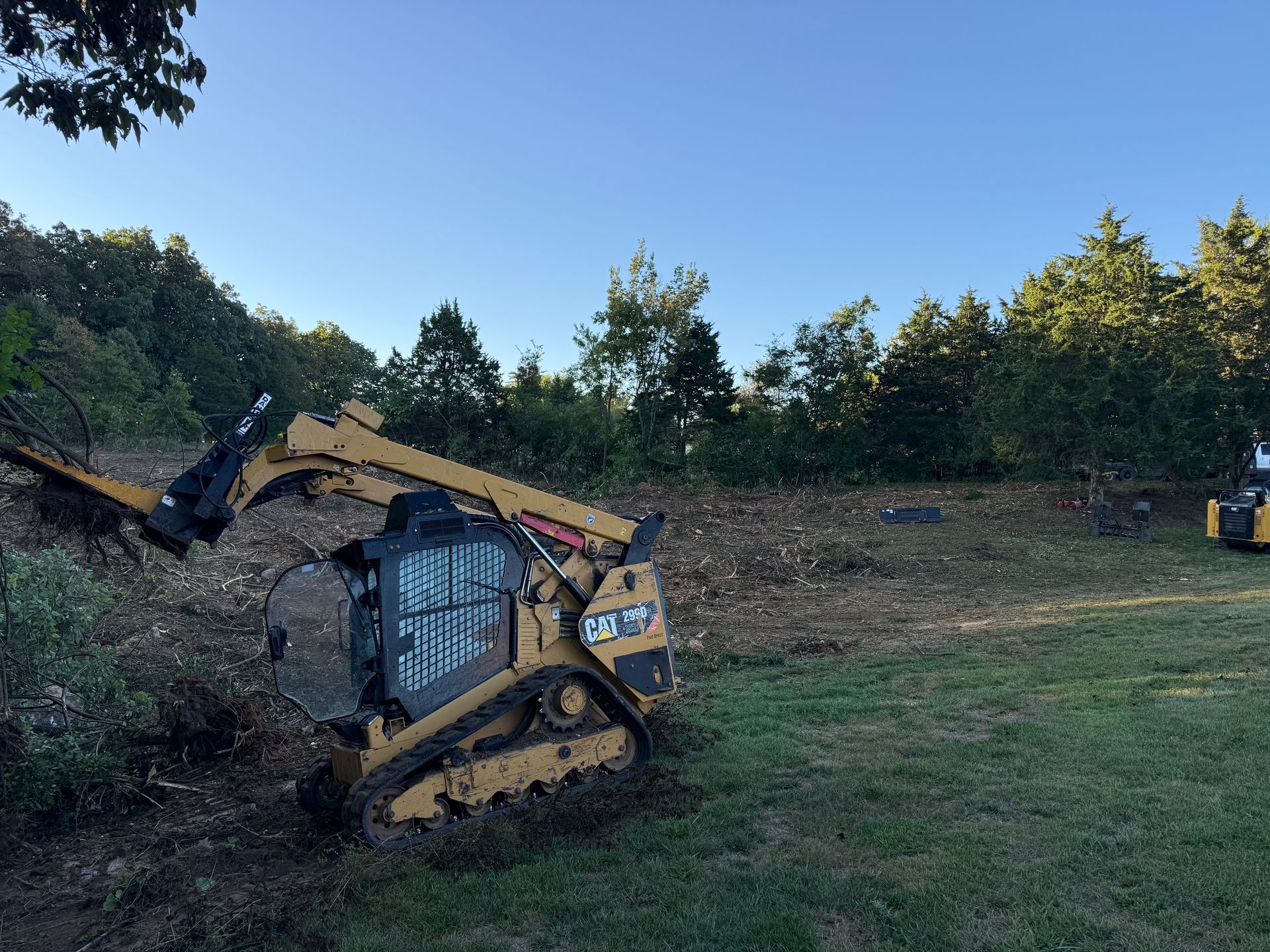 Yellow compact track loader on a grassy dirt slope, with another machine in the background under a blue sky.