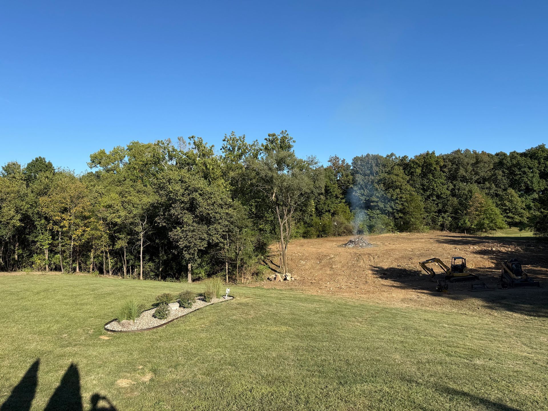 Grassy clearing by trees under a blue sky, with a small smoky fire pit and logs on the right.