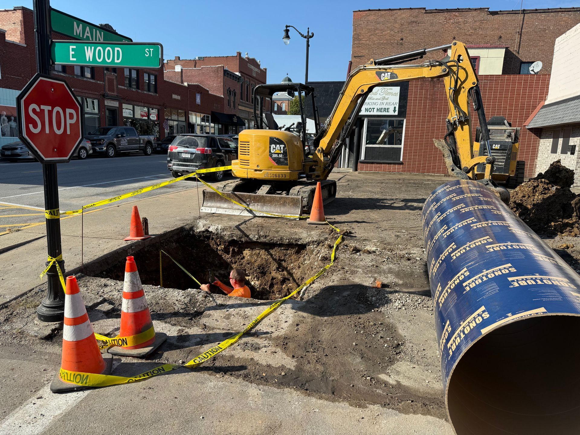 Roadwork at a street corner with open trench, cones, excavator, and large blue pipeline section